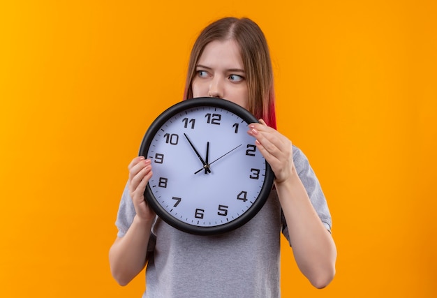 Guardando lato giovane bella ragazza che indossa t-shirt grigia tenendo l'orologio da parete isolato su sfondo giallo