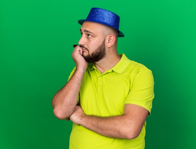 Guardando lato pensando giovane uomo che indossa il cappello da festa blu che tiene il fischio di festa isolato sul muro verde