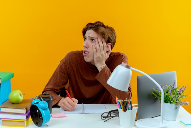 Guardando lato triste giovane studente ragazzo seduto alla scrivania con strumenti di scuola mettendo la mano sulla guancia