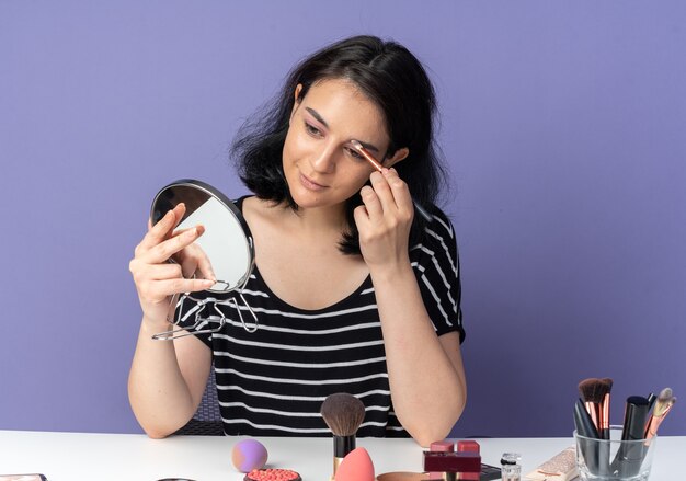 Guardando lo specchio felice dall'aspetto giovane bella ragazza si siede al tavolo con strumenti per il trucco applicando ombretto con pennello per il trucco isolato sulla parete blu