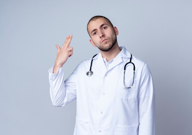 Guardando seriamente il giovane medico maschio che indossa la veste medica e lo stetoscopio intorno al collo che fa il gesto della pistola con la mano isolata sulla parete bianca