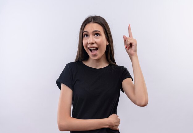 Guardando su gioiosa giovane ragazza caucasica che indossa la maglietta nera punta verso l'alto con il dito su sfondo bianco isolato