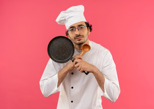 Guardando un giovane cuoco maschio che indossa l'uniforme da chef e gli occhiali che tengono e attraversano la padella con il cucchiaio