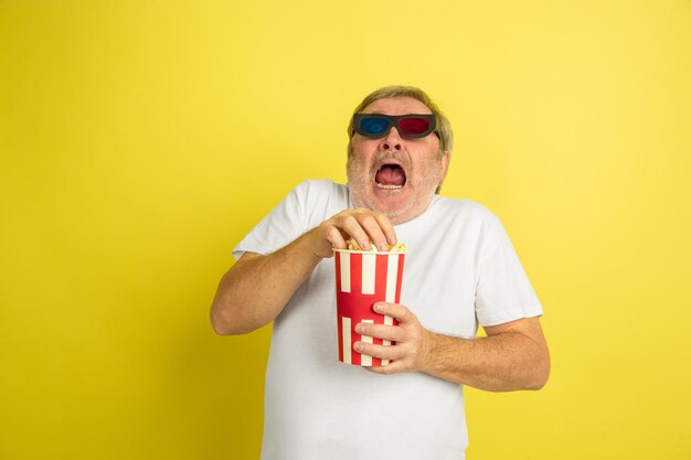 Guardare il cinema con popcorn e occhiali 3D. Ritratto di uomo caucasico su sfondo giallo studio. Bellissimo modello maschile in camicia.