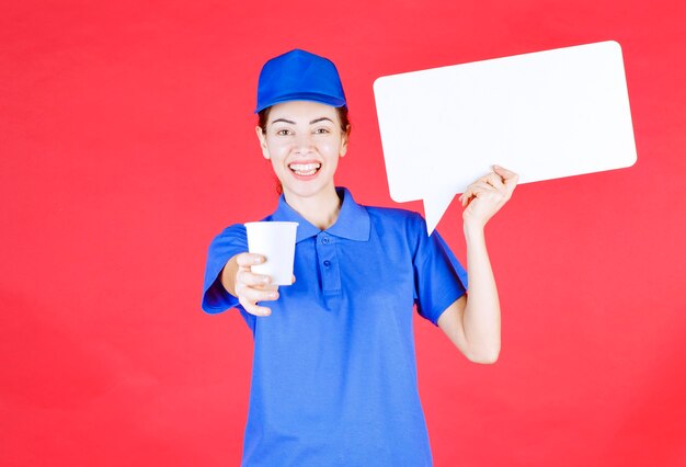 Guida femminile in uniforme blu che tiene in mano un pannello informativo rettangolare bianco e offre una tazza di bevanda usa e getta al partecipante.