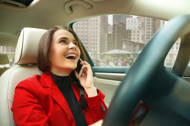 Guidare in città. Giovane donna attraente alla guida di un'auto. Giovane modello abbastanza caucasico in elegante giacca rossa alla moda che si siede all'interno del veicolo moderno