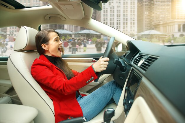 Guidare in città. Giovane donna attraente alla guida di un'auto. Giovane modello piuttosto caucasico in elegante giacca rossa alla moda che si siede all'interno del veicolo moderno. Concetto di imprenditrice.