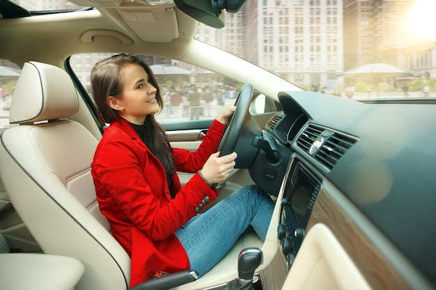 Guidare in città. Giovane donna attraente alla guida di un'auto. Giovane modello piuttosto caucasico in elegante giacca rossa alla moda che si siede all'interno del veicolo moderno