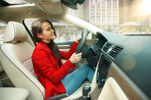 Guidare in città. Giovane donna attraente alla guida di un'auto. Giovane modello piuttosto caucasico in elegante giacca rossa alla moda che si siede all'interno del veicolo moderno