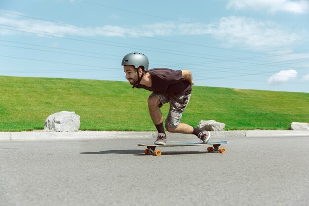 Guidatore di skateboard facendo un trucco in strada nella giornata di sole. Giovane uomo in attrezzatura equitazione e longboard vicino a prato in azione. Concetto di attività per il tempo libero, sport, estremo, hobby e movimento.