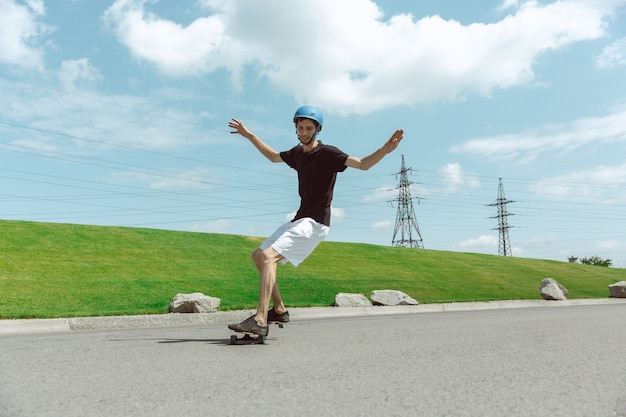 Guidatore di skateboard facendo un trucco sulla strada della città in una giornata di sole. Giovane uomo in attrezzatura equitazione e longboard vicino a prato in azione. Concetto di attività per il tempo libero, sport, estremo, hobby e movimento.