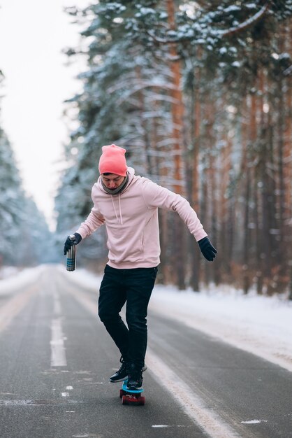 Guidatore di skateboard in piedi sulla strada in mezzo alla foresta circondata dalla neve