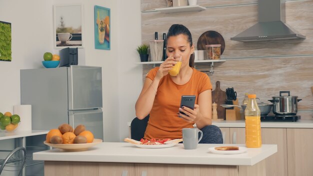 Gustare un bicchiere di succo d'arancia fresco durante la colazione e navigare sullo smartphone. Donna che beve succo d'arancia sano e naturale. Casalinga che beve succo d'arancia sano, naturale e fatto in casa. ricaricare