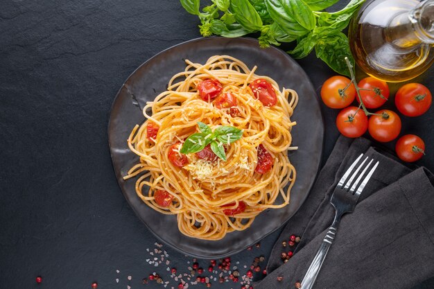 Gustosi e appetitosi spaghetti classici italiani con salsa di pomodoro, parmigiano e basilico sul piatto e ingredienti per cucinare la pasta sul tavolo scuro. Copia spazio piatto vista dall'alto.
