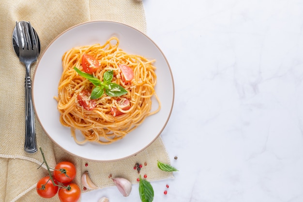 Gustosi e appetitosi spaghetti italiani classici con salsa di pomodoro, parmigiano e basilico sul piatto e ingredienti per cucinare la pasta sul tavolo di marmo bianco. Spazio di copia vista dall'alto piatto.
