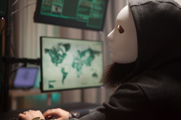 Hacker barbuto con una felpa con cappuccio che indossa una maschera bianca usando il suo computer con più schermi.