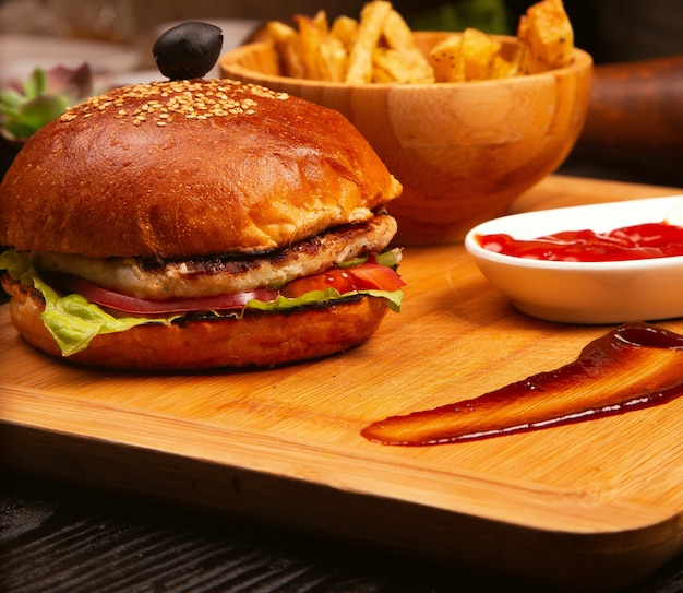 Hamburger di carne di pollo con pomodoro e lattuga all&#39;interno e patatine fritte servite con olive nere e ketchup su un vassoio di legno