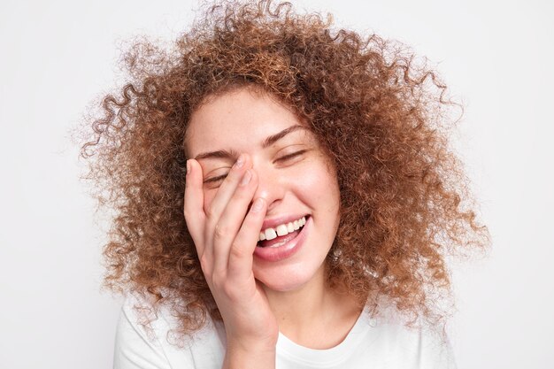 Headshot di bella donna europea sincera con capelli folti ricci si diverte a ridere copre il viso con sorrisi di palme esprime ampiamente la felicità isolata sul muro bianco. Emozioni positive