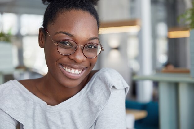 Headshot di femmina africana dalla pelle scura attraente positiva in occhiali rotondi, esprime piacevoli emozioni
