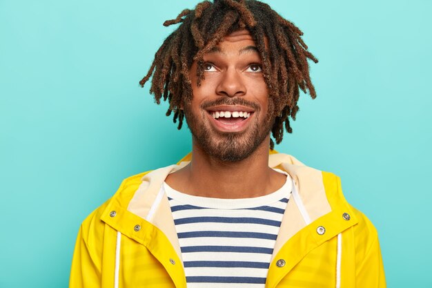 Headshot di uomo felice con dreadlocks, ha un'espressione positiva, focalizzata sopra, indossa un maglione da marinaio e un impermeabile giallo, isolato su sfondo blu