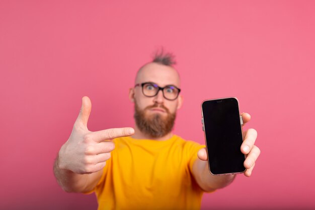 Hey qualcosa di nuovo. Ragazzo barbuto europeo arrabbiato serio che indica il suo telefono cellulare con lo schermo in bianco nero sul colore rosa