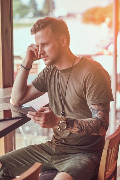 Hipster alla moda bello si siede a un tavolo in un caffè lungo la strada, tiene lo smartphone.