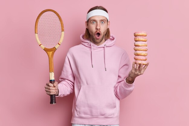 Hipster barbuto scioccato indossa abbigliamento sportivo tiene racchetta da tennis e una pila di ciambelle dolci guarda sorprendentemente con la bocca ampiamente aperta per il tempo libero attivo.