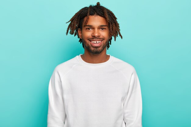 Hipster con i dreadlock, sorride ampiamente, mostra i denti, indossa un maglione casual bianco, soddisfatto dopo aver trascorso il fine settimana con gli amici