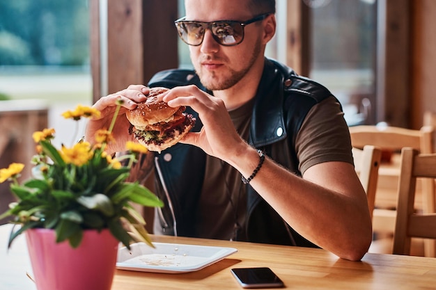 Hipster con un taglio di capelli e una barba alla moda si siede a un tavolo, ha deciso di cenare in un bar lungo la strada, mangiando un hamburger.