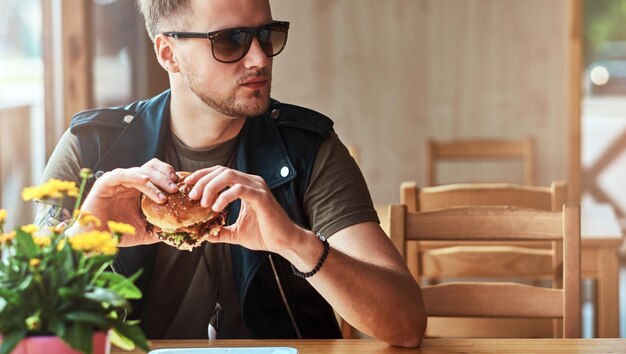 Hipster con un taglio di capelli e una barba alla moda si siede a un tavolo, ha deciso di cenare in un bar lungo la strada, mangiando un hamburger.