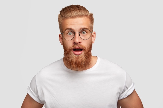 Hipster emotivo con espressione stupita, si chiede le ultime notizie, ha una folta barba rossa e baffi