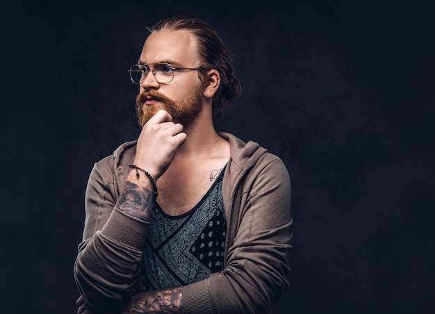 Hipster rosso pensieroso intelligente con barba piena e occhiali vestito con abiti casual, posa con la mano sul mento in uno studio. Isolato su sfondo scuro.