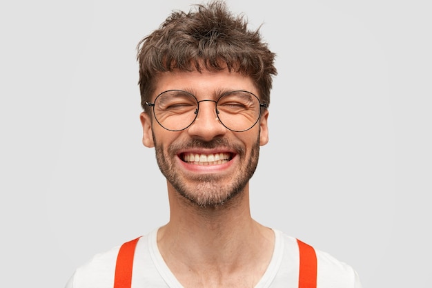 Hipster uomo barbuto positivo sorride ampiamente, ha un'espressione soddisfatta, ride di qualcosa di divertente, chiude gli occhi,
