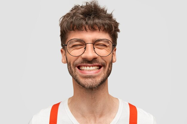 Hipster uomo barbuto positivo sorride ampiamente, ha un'espressione soddisfatta, ride di qualcosa di divertente, chiude gli occhi,