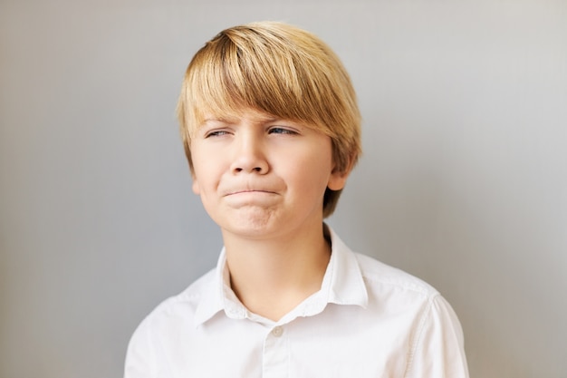 Hmm, fammi pensare. Foto di ragazzo bello emotivo con capelli biondi che perseguono le labbra con espressione facciale pensierosa pensierosa, cercando di ricordare qualcosa, vestito con una camicia bianca, in posa isolato