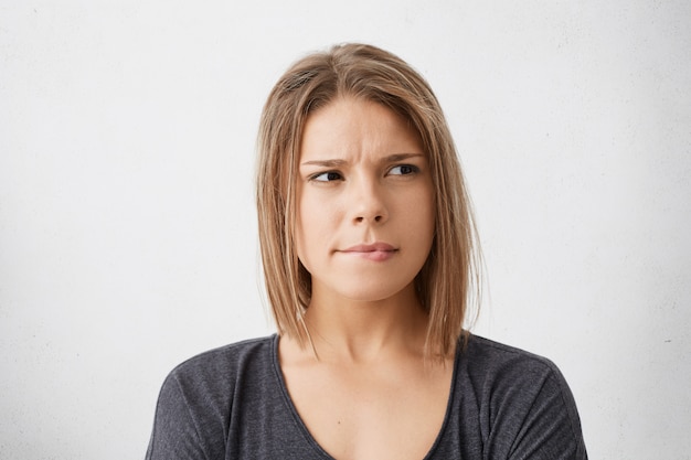 Hmm. Primo piano del volto di dubbiosa carina giovane donna vestita con indifferenza che insegue le labbra, assorta nel pensiero, cercando di trovare la giusta soluzione mentre si tratta di qualche problema, con sguardo indeciso concentrato