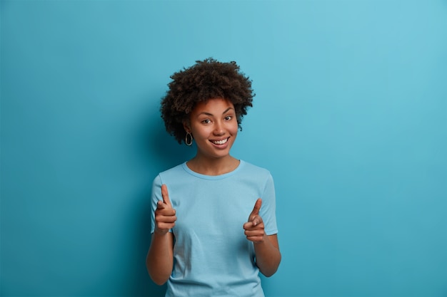 Ho fiducia in te. La ragazza dalla pelle scura positiva fa il gesto della pistola con le dita, incoraggia qualcuno, sceglie di unirsi alla squadra, elogia per un buon lavoro, sorride ampiamente, indossa una maglietta blu casual, sta al coperto