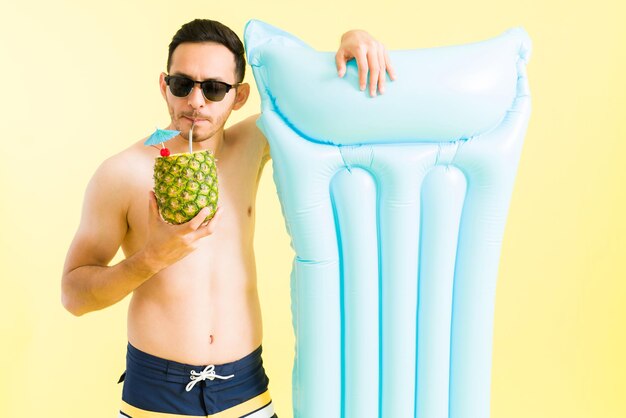 Ho tutto ciò di cui ho bisogno per una vacanza rilassante. Uomo bello che indossa occhiali da sole e beve una pina colada fredda mentre tiene un gonfiabile a bordo piscina