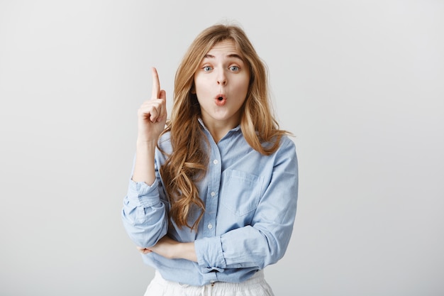 Ho una grande idea. Tiro al coperto di intelligente modello femminile di bell'aspetto con i capelli biondi in camicia da colletto blu che alza il dito indice, dicendo wow mentre si ha un piano o si prepara un suggerimento sul muro grigio