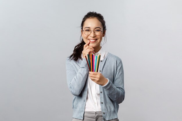 Hobby, creatività e concetto di arte. Sciocco donna asiatica sorridente felice, artista con gli occhiali ridacchia mentre tiene in mano matite colorate, ha un'idea interessante per la sua prossima opera d'arte, disegno, sfondo grigio.