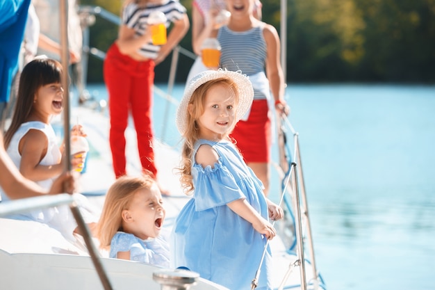 I bambini a bordo dello yacht del mare bevono succo d'arancia. Le ragazze adolescenti o bambini contro il cielo blu all'aperto. Vestiti colorati. Concetti di moda per bambini, estate soleggiata, fiume e vacanze.