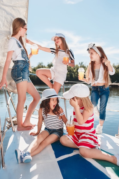 I bambini a bordo dello yacht del mare bevono succo d'arancia. Le ragazze adolescenti o bambini contro il cielo blu all'aperto. Vestiti colorati. Concetti di moda per bambini, estate soleggiata, fiume e vacanze.