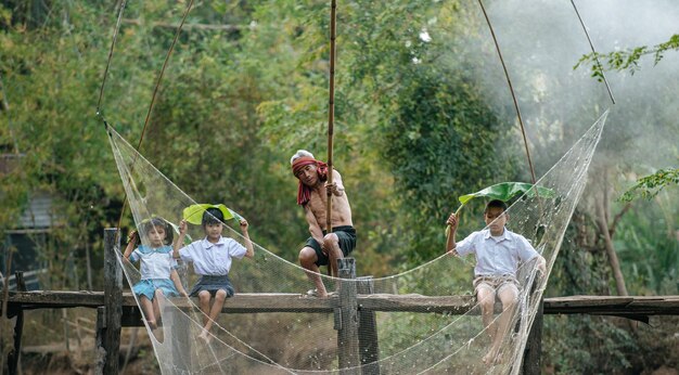 I bambini asiatici si divertono a rallegrare il nonno o il pescatore anziano usano la rete quadrata per catturare il pesce nel canale, tre fratelli allegri sul ponte di legno dopo la scuola, lo spazio della copia, il concetto di stile di vita rurale