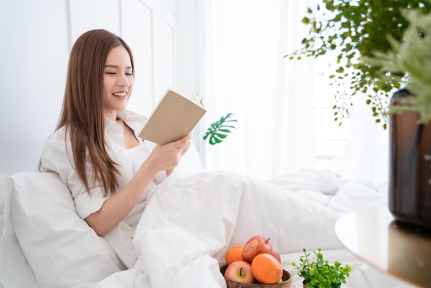 I capelli lunghi del vestito bianco femminile asiatico di bellezza hanno letto il libro sul letto bianco con il fondo interno bianco allegro della camera da letto del mollusco