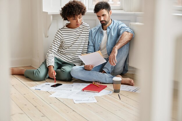 I colleghi interrazziali leggono la richiesta scritta per la produzione di più beni, studiano il prezzo di rivendita seduti insieme sul pavimento a casa