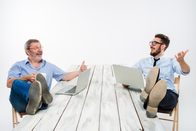I due uomini d'affari sorridenti con le gambe sul tavolo che lavorano su laptop su sfondo bianco. Affari in stile americano