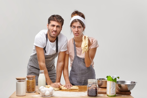 I fornai arrabbiati impastano insieme la pasta, hanno fatto qualcosa di sbagliato, non hanno seguito la ricetta, hanno preparato la pizza fatta in casa, sono sporchi di farina, isolati sul muro bianco, circondati dai prodotti alimentari necessari