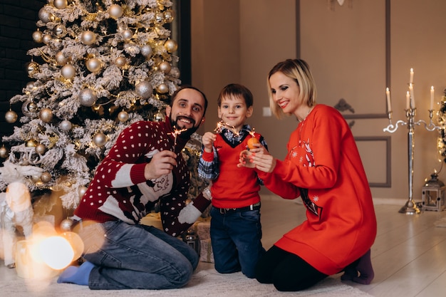 I genitori e il loro figlioletto in maglione rosso si divertono con le arance sedute davanti a un albero di Natale