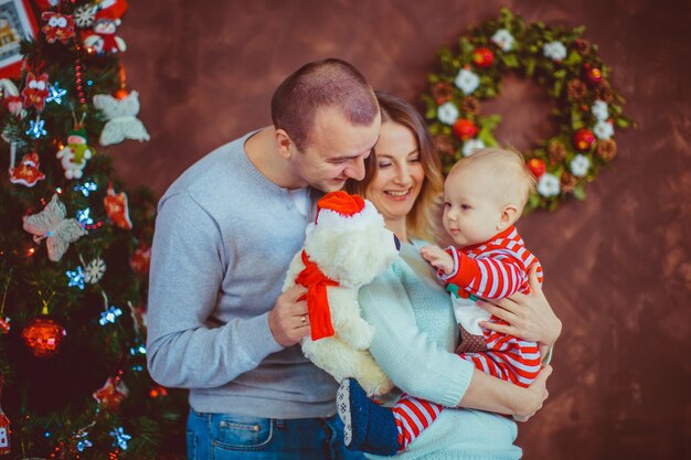 I giovani genitori allegri tengono il piccolo bambino che posa prima di un albero di Natale