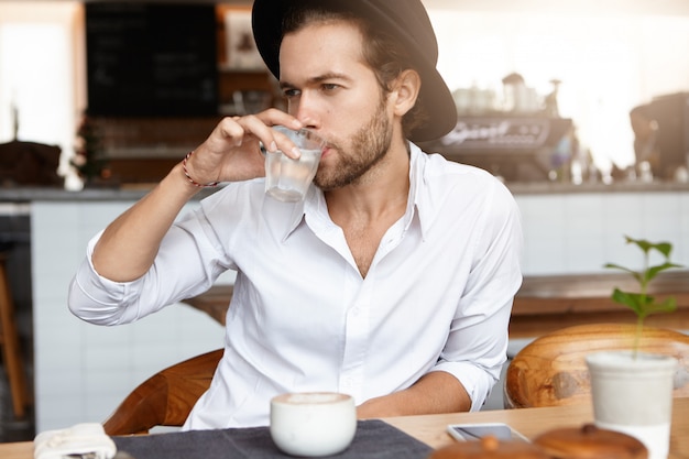 I giovani pantaloni a vita bassa caucasici si sono vestiti in acqua bianca della camicia bianca da vetro durante la pausa caffè al self-service. Uomo barbuto alla moda in black hat che si rilassa da solo nell'interno moderno del caffè. Orizzontale